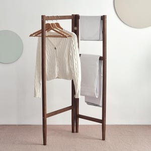 Może przedstawiać: Ciemnobrązowy drewniany wieszak na ubrania z białym kardiganem w warkocze na drewnianych wieszakach. Dwa białe ręczniki są zawieszone na wieszaku. Wieszak jest wolnostojący i ma konstrukcję przypominającą drabinę.