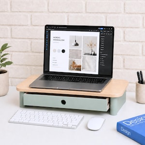Puede incluir: Un soporte de madera para portátil con un cajón verde claro y una parte superior de madera natural. Un portátil muestra el sitio web con el texto "NORDIC MINIMALISM". Un teclado y un ratón blancos están delante del soporte. Un libro azul y un portalápices están a la derecha.