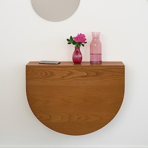 May include: A semi-circular wooden wall-mounted console table with a visible grain pattern. On the flat top surface sits a small pink vase containing a pink flower, a clear glass carafe, and a thin rectangular electronic device.