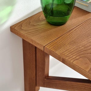 May include: A wooden wall-mounted shelf with a triangular design. The shelf is made of light brown wood and has a smooth finish. A green glass vase is sitting on the shelf.