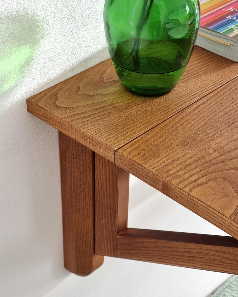 May include: A wooden wall-mounted shelf with a triangular design. The shelf is made of light brown wood and has a smooth finish. A green glass vase is sitting on the shelf.