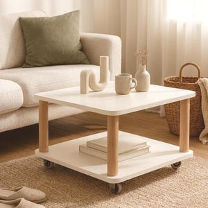 Może przedstawiać: Biały, dwupoziomowy kwadratowy stolik kawowy z jasnymi drewnianymi nogami i kółkami. Na górnej półce znajdują się dekoracyjne wazony i kubek. Na dolnej półce znajdują się książki. Stół stoi na tkanej macie, z sofą i poduszką w tle.