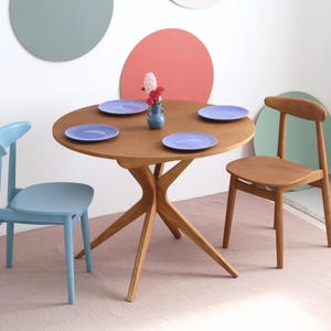 Może przedstawiać: Okrągły drewniany stół jadalny z czterema niebieskimi talerzami, małym niebieskim wazonem z kwiatami i dwoma drewnianymi krzesłami. Widoczne są dwa dodatkowe niebieskie krzesła. Stół ma unikalny kształt podstawy. Tło przedstawia kolorowe okrągłe dekoracje ścienne.
