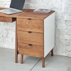 Könnte beinhalten: Ein Schreibtischmodul aus Holz mit drei Schubladen, schwarzen rechteckigen Griffen, einer weißen Seitenwand und vier konischen Holzbeinen. Auf der Oberfläche stehen ein silberner Laptop und ein Buch. Die Holzmaserung ist an den Fronten sichtbar.