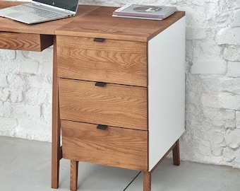 May include: A wooden desk unit featuring three drawers with black rectangular handles, a white side panel, and four tapered wooden legs. The top surface holds a silver laptop and a thin book. The wood grain is visible on the drawer fronts.