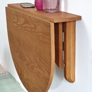 May include: A wall-mounted, semi-circular wooden drop-leaf table with a natural grain finish. The table features a hinged mechanism that allows the curved leaf to fold downward against the wall, supported by a vertical wooden bracket.