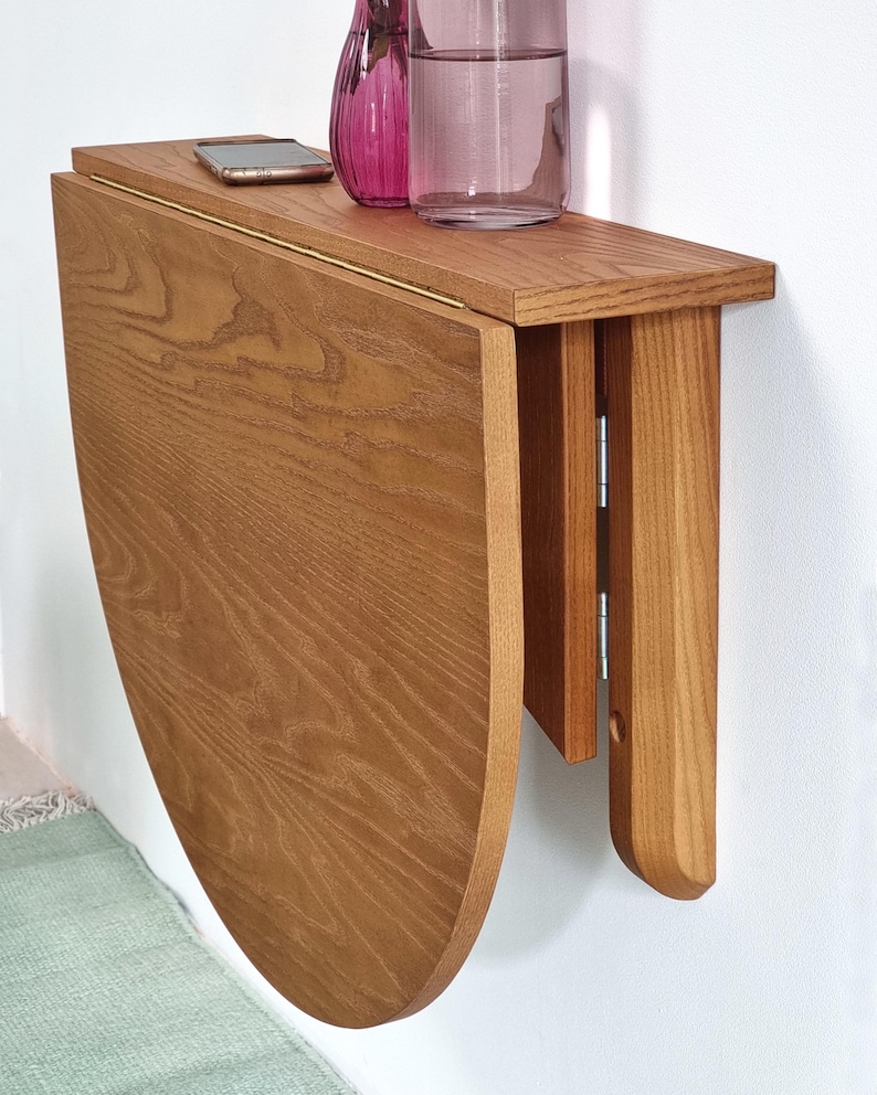 May include: A wall-mounted, semi-circular wooden drop-leaf table with a natural grain finish. The table features a hinged mechanism that allows the curved leaf to fold downward against the wall, supported by a vertical wooden bracket.