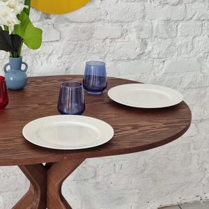 May include: A wooden oval table with a dark brown finish and a cross-shaped base. Two white plates and two blue glasses are on the table.