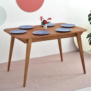Może przedstawiać: Drewniany stół jadalny z sześcioma niebieskimi talerzami i małym wazonem z czerwonymi kwiatami. Stół ma prostokątny blat i skośne nogi. Talerze są ułożone na stole, gotowe do posiłku.