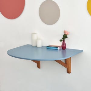 Könnte beinhalten: Ein hellblaues, halbkreisförmiges Wandregal mit Holzstützen. Auf dem Tisch stehen zwei weiße Kerzen, ein Buch und eine kleine rosa Vase mit einer Rose. Dekorative Kreise in Rot, Grau und Gelb zieren die weiße Wand.