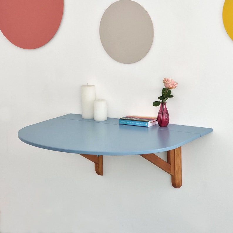 May include: A light blue, semi-circular wall-mounted table with wooden supports. The table holds two white candles, a book, and a small pink vase with a rose. Decorative circles in red, gray, and yellow adorn the white wall.
