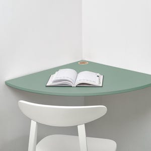 May include: A sage green corner desk with a small hole and an open book. A white chair is positioned below the desk. The desk is mounted on a white wall. The desk is a triangular shape.