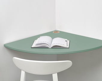 May include: A sage green corner desk shelf mounted against white walls, featuring a curved front edge and a circular cutout for cable management. An open white planner with black text and grid lines rests flat on the smooth, matte surface.