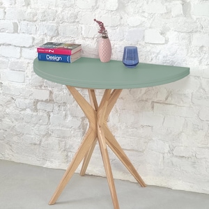 Può includere: Un tavolo a forma di mezzaluna con un piano verde chiaro e una base in legno con quattro gambe. Il tavolo è decorato con un vaso di fiori rosa e un bicchiere d'acqua.