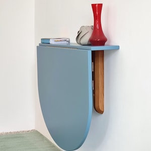 May include: A wall-mounted, light blue, folding table with a wooden support. A red vase, a gray decorative object, and a small stack of books rest on the table's surface. The table is mounted on a white wall.