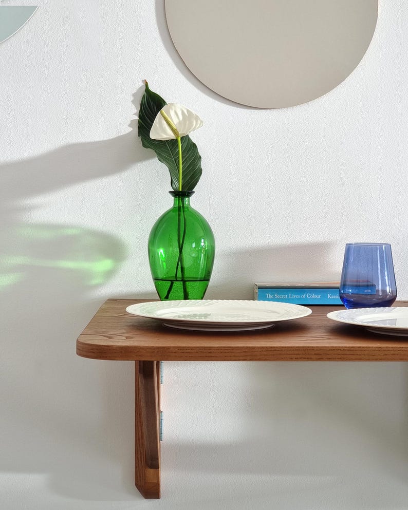 May include: A wooden shelf with a green glass vase holding a white flower and a green leaf. There is a white plate, a blue glass, and a book titled "The Secret Lives of Colour" on the shelf.