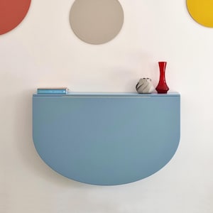 May include: A light blue, semi-circular wall-mounted shelf. A small book rests on the shelf's left side, with a decorative gray and white vase and a red vase on the right. The shelf is mounted on a white wall with colorful circular decorations.