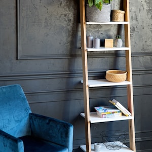 Op de afbeelding: Een witte ladderplank met houten zijkanten, met een potplant, kaarsen, boeken en decoratieve kommen. De plank staat tegen een donkergrijze muur met witte lijstwerk.