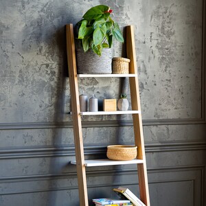 Op de afbeelding: Een houten ladderrek met witte planken, met een potplant, kaarsen, een kleine kom en boeken. Het rek staat tegen een grijze, getextureerde muur.