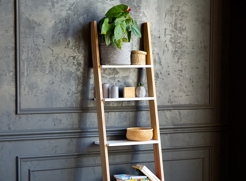 Op de afbeelding: Een houten ladderrek met witte planken, met een potplant, kaarsen, een kleine kom en boeken. Het rek staat tegen een grijze, getextureerde muur.