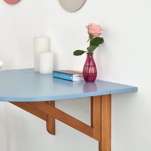 May include: A light blue, wall-mounted, half-circle table with wooden supports. A pink rose in a small, pink vase sits on the table, along with a book and two white candles. Two decorative wall hangings are visible in the background.