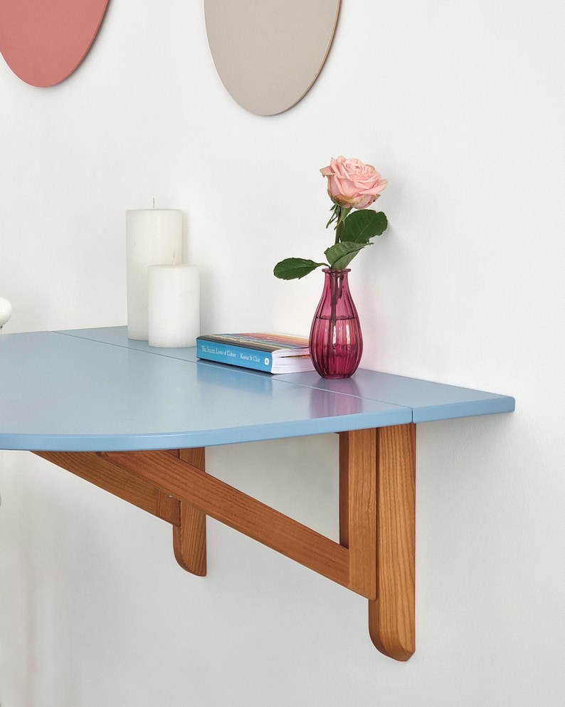 May include: A light blue, wall-mounted, half-circle table with wooden supports. A pink rose in a small, pink vase sits on the table, along with a book and two white candles. Two decorative wall hangings are visible in the background.