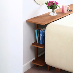 Pode incluir: Uma mesa consola de madeira alta e estreita com um acabamento castanho liso, com três prateleiras abertas. A superfície superior contém um vaso de vidro transparente com flores cor-de-rosa e uma caneca branca com riscas vermelhas verticais.