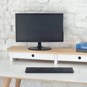 Op de afbeelding: Een zwarte computermonitor staat op een houten monitorstandaard met witte lades. Een zwart toetsenbord staat voor de standaard. Twee boeken liggen aan de rechterkant van de standaard. Het bovenste boek heeft het woord "Design" op de kaft.