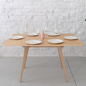 May include: A light wood dining table with a white top and four legs. The table is set with six white plates and six clear glasses. A pink ceramic pineapple sits in the centre of the table.