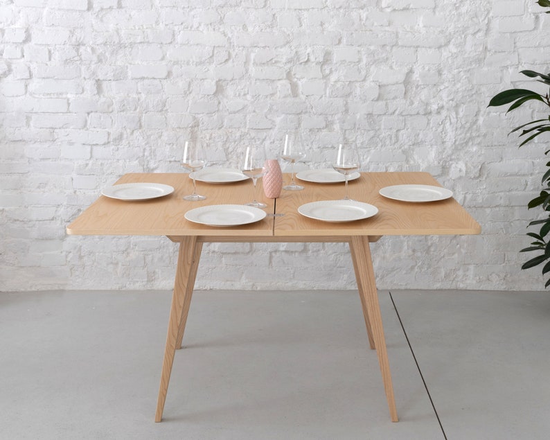 May include: A light wood dining table with a white top and four legs. The table is set with six white plates and six clear glasses. A pink ceramic pineapple sits in the centre of the table.