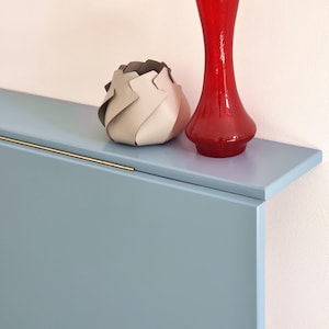 May include: A red glass vase and a decorative, layered, beige and brown object sit on a light blue shelf. The shelf has a gold-colored handle. The background is a light pink wall. The objects are decorative.