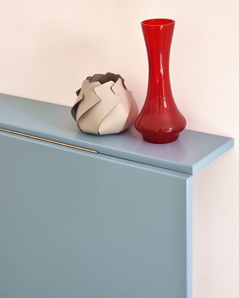 May include: A red glass vase and a decorative, layered, beige and brown object sit on a light blue shelf. The shelf has a gold-colored handle. The background is a light pink wall. The objects are decorative.
