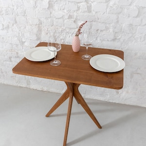 Puede incluir: Pequeña mesa cuadrada de madera marrón cálido. Dos platos blancos, dos copas de vino y un jarrón rosa con flores secas están sobre ella. La mesa tiene una base única de tres patas. Fondo: pared de ladrillo blanco.