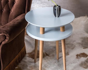 May include: A light blue two-tiered side table with a round top and wooden legs. A white flower in a black and white striped vase sits on the top tier.