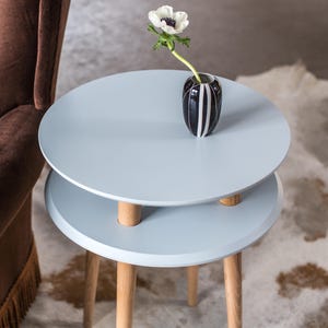 Puede incluir: Una mesa auxiliar redonda de color azul claro con dos niveles y cuatro patas de madera. Un jarr&oacute;n de rayas blancas y negras con una sola flor blanca se encuentra en el nivel superior.