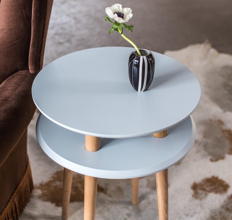Puede incluir: Una mesa auxiliar redonda de color azul claro con dos niveles y cuatro patas de madera. Un jarr&oacute;n de rayas blancas y negras con una sola flor blanca se encuentra en el nivel superior.