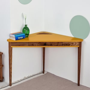 Op de afbeelding: Een driehoekig hoekbureau met een mosterdgeel blad, een ladefront met donkere houtnerf en drie taps toelopende donkere houten poten. Op het bureau staat een stapel boeken met het woord Design op de rug en een groene glazen vaas met een witte bloem.
