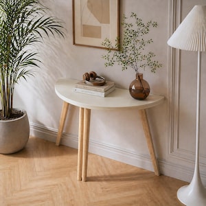 Pode incluir: Uma mesa consola semicircular com um tampo liso em tom branco sujo e três pernas de madeira clara. A mesa sustenta uma pilha de dois livros, um par de auscultadores castanhos e um vaso redondo de vidro âmbar com um ramo verde.
