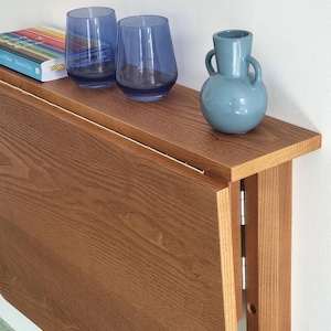 May include: A brown wooden folding wall table with a light wood finish. The table is folded up against a white wall. Two blue glass cups and a blue ceramic vase are on the table's surface.