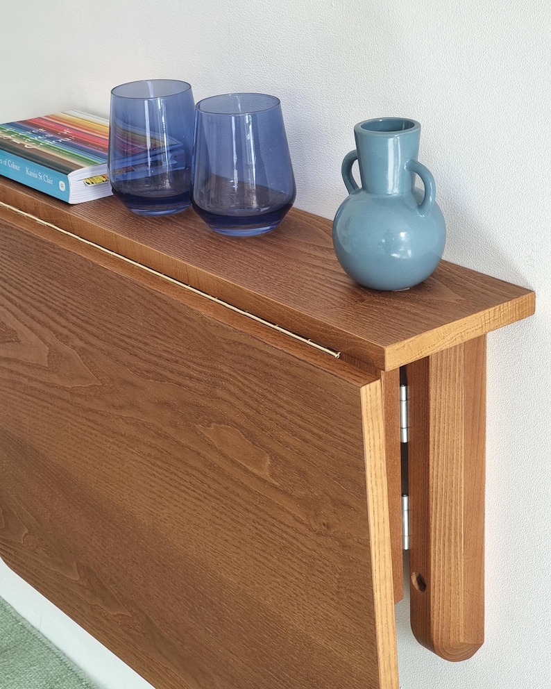 May include: A brown wooden folding wall table with a light wood finish. The table is folded up against a white wall. Two blue glass cups and a blue ceramic vase are on the table's surface.