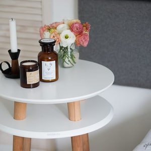 Puede incluir: Mesa auxiliar blanca de dos niveles con patas de madera, que incluye un candelero, una vela y una botella de vidrio con una etiqueta. Un ramo de flores rosas y blancas est&aacute; en un jarr&oacute;n de vidrio transparente sobre la mesa.