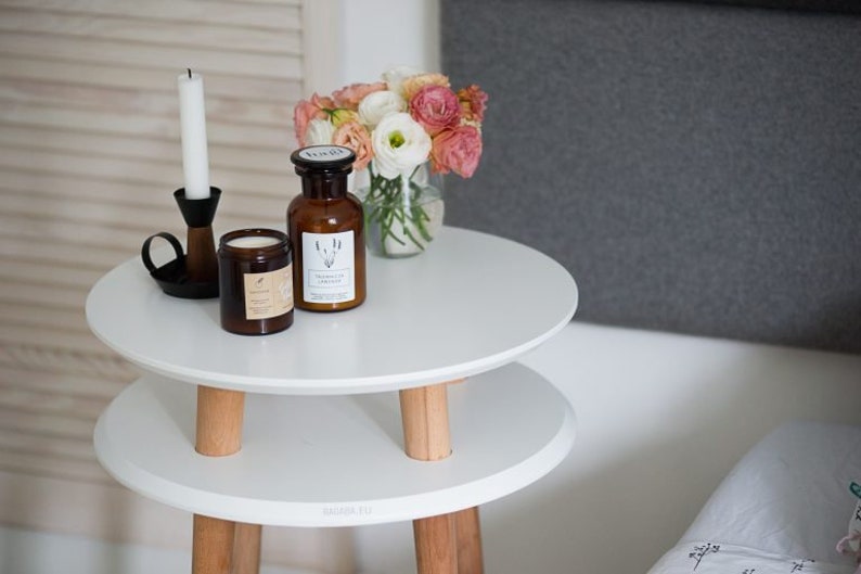 Puede incluir: Mesa auxiliar blanca de dos niveles con patas de madera, que incluye un candelero, una vela y una botella de vidrio con una etiqueta. Un ramo de flores rosas y blancas est&aacute; en un jarr&oacute;n de vidrio transparente sobre la mesa.