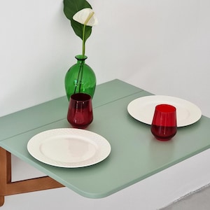 Może przedstawiać: Jasnozielony składany stolik ścienny z dwoma białymi talerzami i dwoma czerwonymi szklankami. Zielony szklany wazon z białym kwiatem stoi na stole.