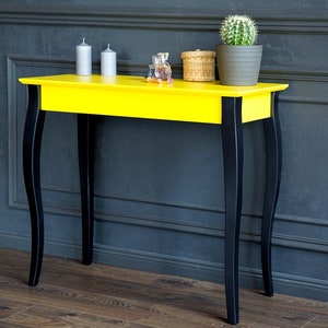 Może przedstawiać: Żółty stolik konsolowy z czarnymi nogami. Stolik ma prosty design z prostokątnym blatem i zakrzywionymi nogami. Stolik stoi przed szarą ścianą.