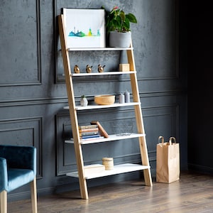 May include: A five-tier ladder shelf with light wood side frames and white horizontal shelves. The shelves hold a framed print, a potted plant, three small bird figurines, a woven bowl, candles, books, a wire basket, and a small lidded container.