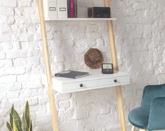 May include: A ladder desk with light wood legs and a white desktop featuring two drawers with black knobs. The unit includes an upper shelf holding white binders, books, a radio, and a plant. A teal velvet chair sits nearby.
