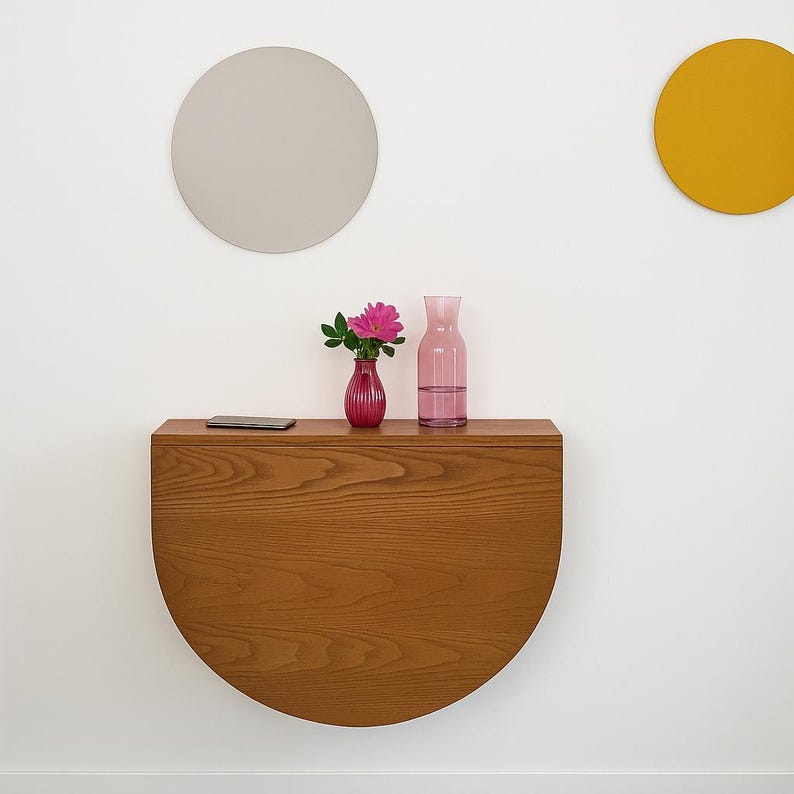 May include: A semi-circular wooden wall-mounted console table with a visible grain pattern. On the flat top surface sits a small pink vase containing a pink flower, a clear glass carafe, and a thin rectangular electronic device.