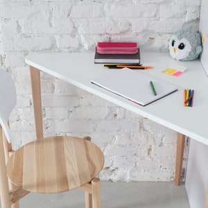 Può includere: Una scrivania triangolare bianca con una cornice in legno si trova in un angolo contro un muro di mattoni bianchi. Una sedia con una seduta in legno chiaro e uno schienale bianco è posizionata accanto alla scrivania. La scrivania è decorata con materiale scolastico, tra cui un quaderno, matite, pastelli e una custodia per matite rosa.