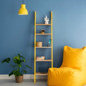 Puede incluir: Estantería amarilla en forma de escalera con estantes de madera contra una pared azul. Los estantes exhiben un cactus blanco, un reloj, una cesta tejida, libros y una cesta de alambre negra. Un puf amarillo está cerca.