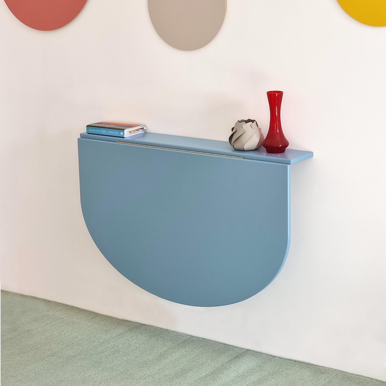 May include: A light blue, semi-circular wall-mounted table with a shelf. A small book, a white and gray decorative object, and a red vase are on the shelf. The table is attached to a white wall.